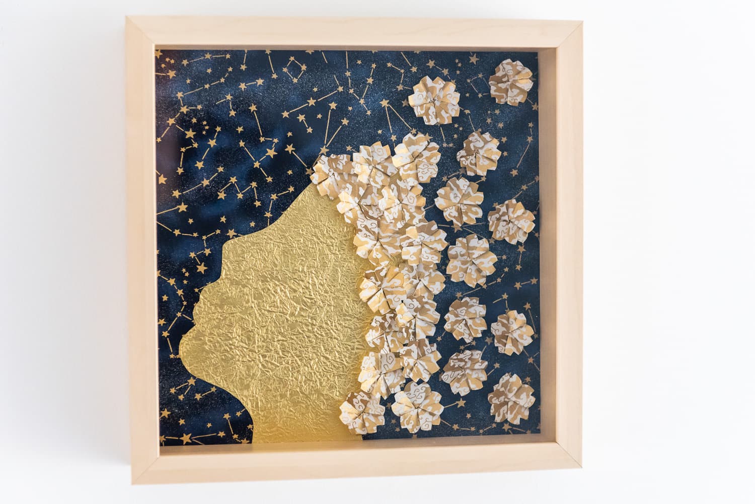 Cadre mural doré représentant un profil de visage sur fond étoilé, avec des fleurs en papier washi doré et blanc.